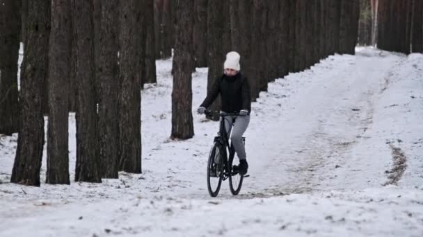 若い女性は 松林の冬の木の間の雪の道で自転車に乗る スローモーション ウッドランドトラックで自転車に乗るサイクリストの1人 自転車で自転車に乗る 健康的なライフスタイル 積極的な趣味 スポーツ — ストック動画
