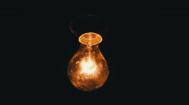 Hanging incandescent bulb turns on, and off and flickers on black background, place for text. Warm flashing filament close-up. Glass old bulb glowing yellow warm light. Tungsten lamp light. Idea 4K