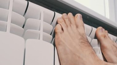 Erkek çıplak ayaklar evdeki ısıtıcı kalorifere dokunarak ısınmaya çalışır. Kış mevsiminde apartmanın ısıtıcı kaloriferlerine yakın çekim donmuş bacaklar. Isıtma sistemi zayıf, sıcaklık yok.