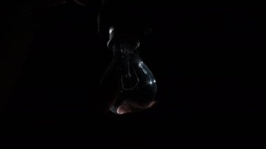 El, ampulü prize çevirir ve siyah arka planda parlar ve titreşir. Isınan filament yakın plan. Camdan yapılmış, sarı, parlak bir ampul. Tungsten lamba ışığı