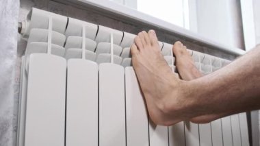 Erkek çıplak ayaklar evdeki ısıtıcı kalorifere dokunarak ısınmaya çalışır. Kış mevsiminde apartmanın ısıtıcı kaloriferlerine yakın çekim donmuş bacaklar. Isıtma sistemi zayıf, sıcaklık yok.
