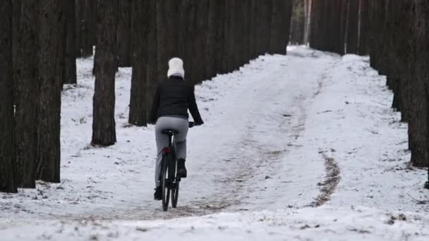 年轻女子骑着自行车在松林中的冬树间的雪地小径上骑着 动作缓慢 一个骑自行车的骑自行车的人骑在林地小径上 骑自行车的人骑自行车 健康的生活方式 积极的爱好 — 图库视频影像