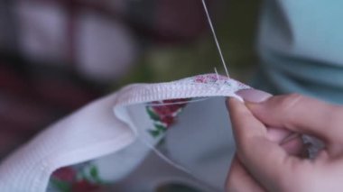 A young woman sews manually with thread and needle in slow motion, close-up. Female fingers with manicures sew while sitting in casual clothes on the sofa at home. Cozy atmosphere, lifestyle, handmade