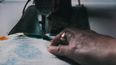 An aged seamstress sews at a traditional sewing machine at home in slow motion. Close-up of aged female hands working at a retro sewing machine. The needle quickly moves up and down. Lifestyle, hobby