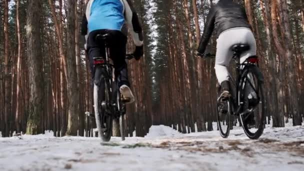2人は一緒にゆっくりとした動きで雪の木の間の冬の道に沿って2台の自転車に乗る 女と男の自転車は田舎道をたどります 2人の自転車が黒い自転車で移動する ライフスタイル スポーツ — ストック動画