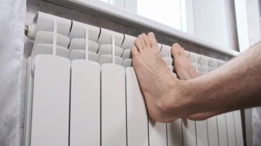 Erkek çıplak ayaklar evdeki ısıtıcı kalorifere dokunarak ısınmaya çalışır. Kış mevsiminde apartmanın ısıtıcı kaloriferlerine yakın çekim donmuş bacaklar. Isıtma sistemi zayıf, sıcaklık yok.