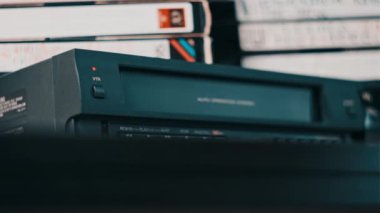 Male hand inserts VHS cassette into a VCR Video recorder. Black vintage videotape cassette recorder on desk with many video cassettes. Inserting retro VHS tape into vintage player. Home video archive