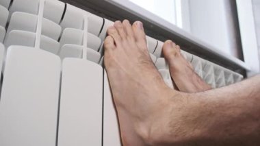 Erkek çıplak ayaklar evdeki ısıtıcı kalorifere dokunarak ısınmaya çalışır. Kış mevsiminde apartmanın ısıtıcı kaloriferlerine yakın çekim donmuş bacaklar. Isıtma sistemi zayıf, sıcaklık yok.