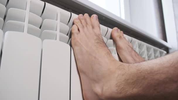 男性の裸の足は家の暖房ラジエーターに触れることによって暖かく保つことを試みる 寒い足は冬の季節にアパートの暖房ラジエーターにクローズアップします 概念悪い暖房システム 暖かさなし — ストック動画
