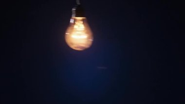 Incandescent lamp on wire sways from side to side and lights up with flickers on dark blue background. Classic bulb turned on and off close-up. Tungsten bulb cozy shine of vintage light. Copy space 4K