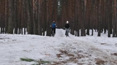 A couple of man with woman ride a bicycle along a snowy path in a winter pine forest, slow motion. Biking couple follow track between snowy trees. Two cyclists travel on black bikes. Lifestyle, sport