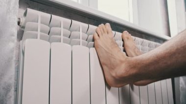 Erkek çıplak ayaklar evdeki ısıtıcı kalorifere dokunarak ısınmaya çalışır. Kış mevsiminde apartmanın ısıtıcı kaloriferlerine yakın çekim donmuş bacaklar. Isıtma sistemi zayıf, sıcaklık yok.