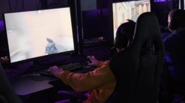 Esport kulübündeki bir genç, oyun sandalyesinde monitörün önünde oturup atış oyunu oynuyor. İnternet kafede online turnuva oynayan çocuklar, neon ışıkları olan profesyonel oyun bilgisayarları..