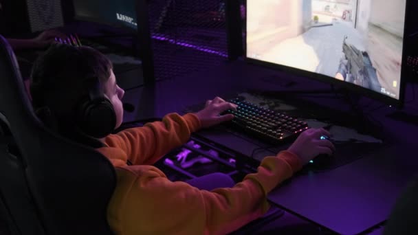 若いゲーマーはプロのゲームチェアに座って Eスポーツクラブでビデオゲームをプレイします ネオンライト付きのコンピュータクラブのヘッドセットの少年は ビデオゲームに焦点を当てています ゲームクラブの10代の若者趣味 — ストック動画