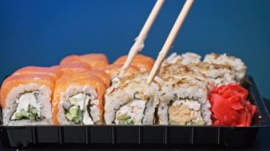 Sopalar suşi kutusundan suşi alır, yakın plan. Plastik bir kutuda yuvarlanıyor. Çeşitli lezzetli Japon suşilerinden oluşan hazır yemek seti teslimat konteynerinde. Sağlıklı beslenme, Asya yemekleri, hazır yemek.