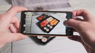 Bir erkek eli akıllı telefon kamerasıyla plastik bir kutuda suşi rulolarının fotoğrafını çeker. Blogcu, sosyal ağlarda yayınlanmak üzere cep telefonundan Japon yemeklerini eleştiriyor. Seçici odak