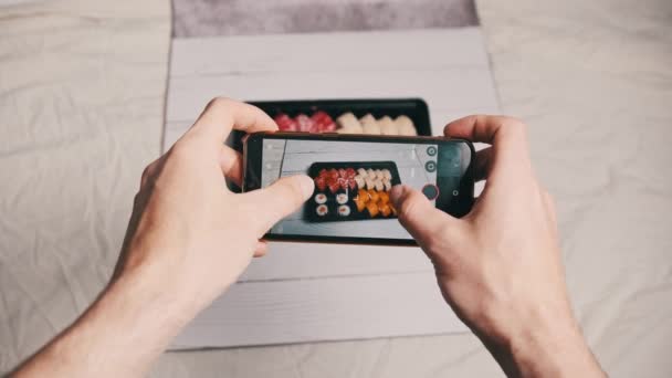 Snsに投稿するためにスマートフォンのカメラで寿司ロールの写真を撮っているブロガー 携帯電話の宅配プラスチック製の箱で日本食を撮影する男性の手 ソーシャルメディアのための撮影 — ストック動画