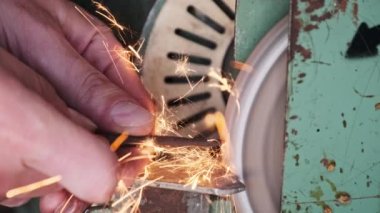 Atölyede aşındırıcı bir tekerlek üzerinde çalışan bir zanaatkarın elleri. Elektrikli öğütme çarkında metal öğütme, yakın plan. Metal bir parçayı incelerken parlak kıvılcımlar çıkıyor. Yavaş çekim