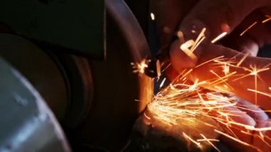 Metal bir parçayı ağır çekimde işlemekten parlak kıvılcımlar çıkıyor. Elektrikli öğütme çarkında metal öğütme, yakın plan. Bir atölyede aşındırıcı bir tekerlek üzerinde çalışan bir zanaatkarın elleri..