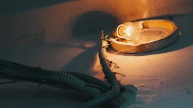 Merdiven boşluğundaki beton duvarda, kırık bir lambanın üzerinde eski bir parlak ampul parlıyor. Filament lambası, duvarda örümcek ağlarıyla sabitlenmiş eski bir elektrik kartuşunda loş bir şekilde parıldıyor..