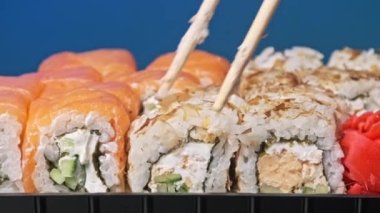 Sopalar suşi kutusundan suşi alır, yakın plan. Plastik bir kutuda yuvarlanıyor. Çeşitli lezzetli Japon suşilerinden oluşan hazır yemek seti teslimat konteynerinde. Sağlıklı beslenme, Asya yemekleri, hazır yemek.