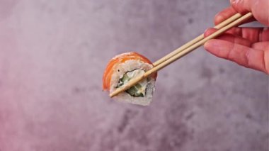 Bulanık arka plandaki gösteriler için suşi çubukları kullanarak bir suşi rulosu al ve tut, kapat. Çubuklu insan eli somon ve Philadelphia peynirli taze Japon suşisi gösteriyor. Deniz ürünleri.