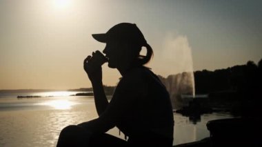 Gün batımında ağır çekimde kahve içen bir kadının silueti. Bir kadın elinde kahveyle nehrin kenarındaki setin kenarında, batan kırmızı güneşin ışınlarında oturuyor. Neşeli, sağlıklı.