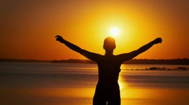 Genç ve rahat bir kadının silueti yavaşça günbatımının tadını çıkarırken ellerini kaldırıyor. Batan güneşe bakan genç bir kızın arka görüntüsü. Yaşam tarzı, özgürlük, mutluluk, zihin
