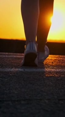 Yavaş çekimde gün batımına doğru yürüyen bir kızın dikey silueti. Hayattan zevk alan narin, atletik bir genç kadının bacaklarının alt görüntüsü. Ruh sağlığı konsepti, yaşam tarzı, özgürlük, mutluluk.