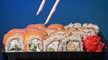 Sopalar suşi kutusundan suşi alır, yakın plan. Plastik bir kutuda yuvarlanıyor. Çeşitli lezzetli Japon suşilerinden oluşan hazır yemek seti teslimat konteynerinde. Sağlıklı beslenme, Asya yemekleri, hazır yemek.
