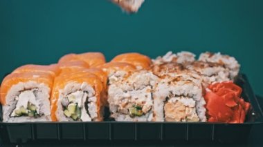 Sopalar suşi kutusuna bir suşi koydu, yakın plan. Plastik bir kutuda yuvarlanıyor. Çeşitli lezzetli suşi setleri teslimat konteynerinde. Sağlıklı beslenme, Asya yemekleri, hazır yemek.