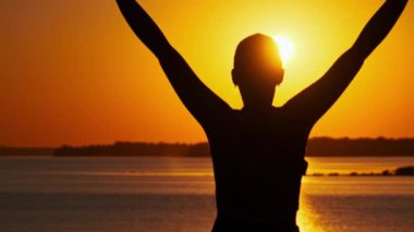 Genç ve rahat bir kadının silueti yavaşça günbatımının tadını çıkarırken ellerini kaldırıyor. Batan güneşe bakan genç bir kızın arka görüntüsü. Yaşam tarzı, özgürlük, mutluluk, zihin