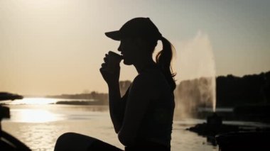 Genç bir kadın güzel bir günbatımında yavaş çekimde sıcak kahve içer. Batan güneşin altındaki kız silueti elinde kahveyle korkulukta oturuyor ve hayatın tadını çıkarıyor. Mutluluk, sağlıklı yaşam tarzı.