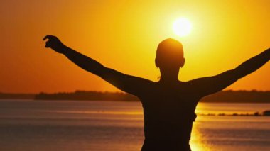 Genç bir kadının silueti ellerini günbatımına doğru kaldırıyor, ağır çekimde. Doğada hayatın tadını çıkaran ve batan güneşe bakan genç bir kız. Özgürlüğü, mutluluğu, akıl sağlığını kavrayın.