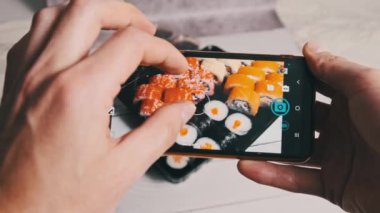 Bir erkek eli akıllı telefon kamerasıyla plastik bir kutuda suşi rulolarının fotoğrafını çeker. Blogcu, sosyal ağlarda yayınlanmak üzere cep telefonundan Japon yemeklerini eleştiriyor. Seçici odak