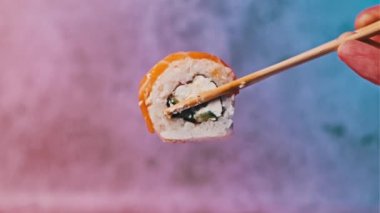 Suşi olarak çubuk kullanarak bir suşi rulosunu al ve götür, yakın plan. Çubuklu insan eli, arka planda somon ve Philadelphia peynirli taze Japon suşisini gösteriyor. Deniz ürünleri.