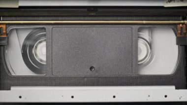 VHS kaset bir VCR kaydedicinin içinde oynatılıyor, üst görünümde. Videokaset makaraları dönüyor. Boş etiketli video kaset oynatmaya başlıyor. İçerideki eski video kaydedici. Klasik film oynatılıyor