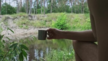 Turist yazın açık havada demir bardakta kahve içiyor. Adam fincanı elinde tutuyor. Nehir kenarındaki yeşil bir zemin üzerinde. Anın tadını çıkarıyor, kamp konsepti, yürüyüş, yakın plan..