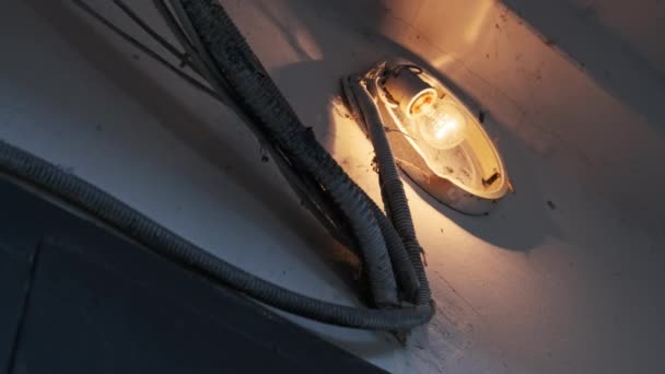一盏白炽灯在水泥墙上的一盏破灯里发出光芒 房子里有危险电线的建筑物照明 楼梯井中有蜘蛛网的旧钨球 — 图库视频影像