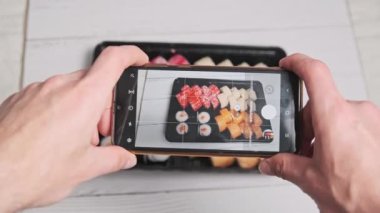 Bir erkek eli akıllı telefon kamerasıyla plastik bir kutuda suşi rulolarının fotoğrafını çeker. Blogcu, sosyal ağlarda yayınlanmak üzere cep telefonundan Japon yemeklerini eleştiriyor. Seçici odak
