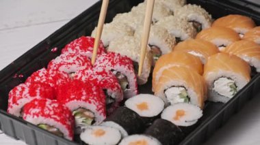 Sopalar suşi kutusundan suşi alır, yakın plan. Plastik bir kutuda yuvarlanıyor. Çeşitli lezzetli Japon suşilerinden oluşan hazır yemek seti teslimat konteynerinde. Sağlıklı beslenme, Asya yemekleri, hazır yemek.