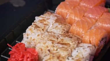 Suşi plastik bir teslimat kutusunda yuvarlanır. Yakın çekim döner. İçinde taze Japon dürümleri, zencefil ve plastik bir kapta dönen wasabi olan suşi kutusu. Yemek teslimatı, paket yemek.