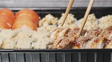 Yemek çubukları suşi kutusundan suşi alır, yakın plan. Plastik bir kutuda yuvarlanıyor. Çeşitli lezzetli Japon suşilerinden oluşan hazır yemek takımı bir teslimat konteynerinde yuvarlanıyor. Yemek teslimatı kavramı.