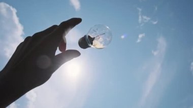 Erkek el tutuşur ve parlak güneşe karşı gökyüzüne parlak bir ampul atar. Şeffaf bir elektrik lambasını tutan insan elinin POV silueti. Ekoloji, güneş enerjisi, eko enerji tasarrufu