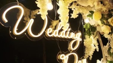 Nikah Günü 'nün neon yazıtları. Düğün kemeri taze beyaz çiçeklerle süslenir ve geceleri ampullerle aydınlatılır. Fotoğraf bölgesi. Pürüzsüz kamera hareketi 4K.