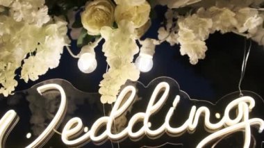 Düğün kemeri. Törende düğün gününün neon yazısıyla birlikte. Tatil kompozisyonu taze beyaz çiçeklerle süslenir ve geceleri ampullerle aydınlatılır. Yumuşak kamera hareketi.