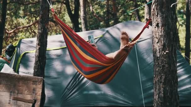 在夏令营现场的吊床上摇晃时使用智能手机的女人 女旅行者在帐篷附近的松树林中使用手机 检查社交网络 生活方式 — 图库视频影像