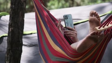 Bir kadın turist çadırın yanındaki hamakta uzanıyor ve akıllı telefon kullanıyor. Dişi ayaklarının görüntüsü. Hamakta sallanan ve yaz kampı yakınlarındaki bir ormanda sosyal medyada sallanan biri. Tatil günü. 4K