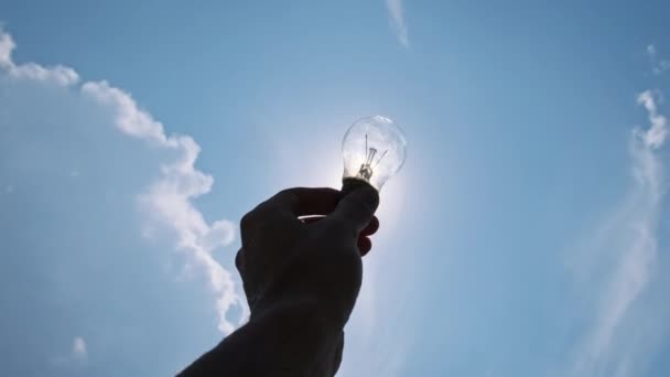 男性の手は青空と明るい太陽に対して白熱電球を保持しています 太陽の光を浴びる透明な電気ランプを握る人間の手のPovシルエット エコロジーコンセプト — ストック動画