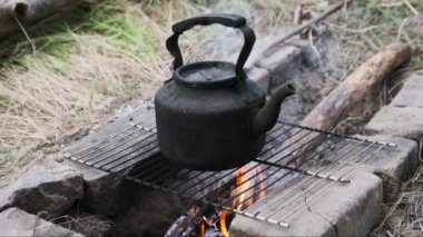Ateşin üstünde çaydanlık. Eski model tütsülenmiş bir çaydanlık, yerdeki taşlardan yapılmış kendi kendini yetiştirmiş turist ocağının üzerinde kaynar su. Kampın yakınındaki ormanda şenlik ateşinde çay yapmak, yakın plan..